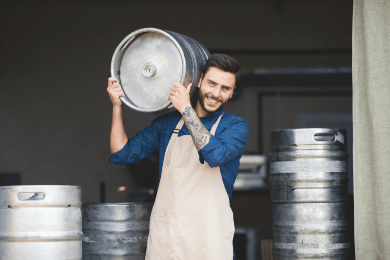 Jonas Leroy smiling effortlessly while carrying a stainless steel beer keg on his shoulder in the brewery warehouse.