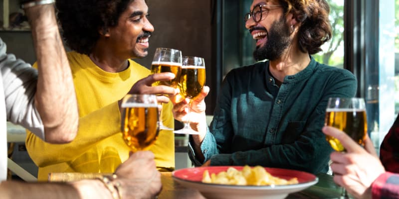 A close-up of a group of friends laughing and clinking glasses of golden beer in a vibrant indoor social setting.