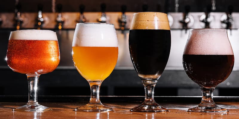 Four different glasses of craft beer, ranging from light pale ale to dark stout, lined up on a bar counter with a row of beer taps in the background.