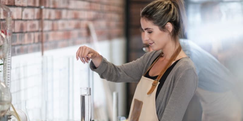 A woman in a brewery apron carefully using a hydrometer to measure the gravity of a clear liquid in a graduated cylinder.