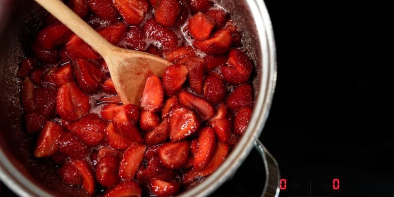 Freshly sliced strawberries simmering in a stainless steel pot with a wooden spoon for a homemade syrup recipe.