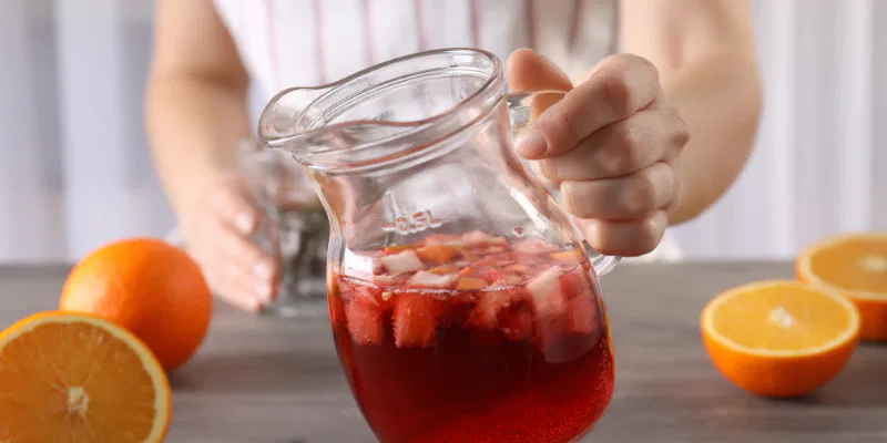 A person holding a glass pitcher filled with a red, fruit-infused beverage surrounded by fresh orange halves.