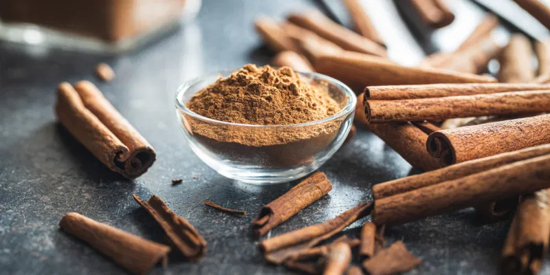 A small glass bowl of ground cinnamon surrounded by whole cinnamon sticks on a dark countertop.