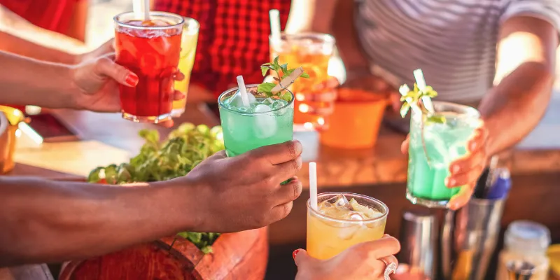 A group of friends cheering with various colorful cocktails and mocktails, including green mojitos and orange fruit drinks, at an outdoor bar.