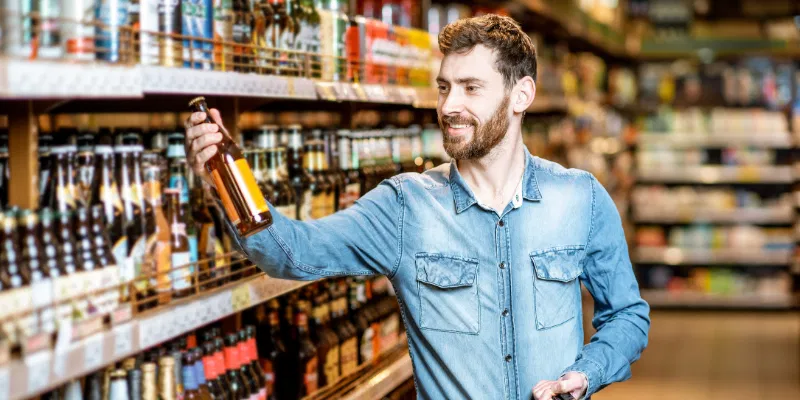 A man in a blue denim shirt standing in a supermarket aisle, holding and inspecting a brown glass beer bottle while shopping.