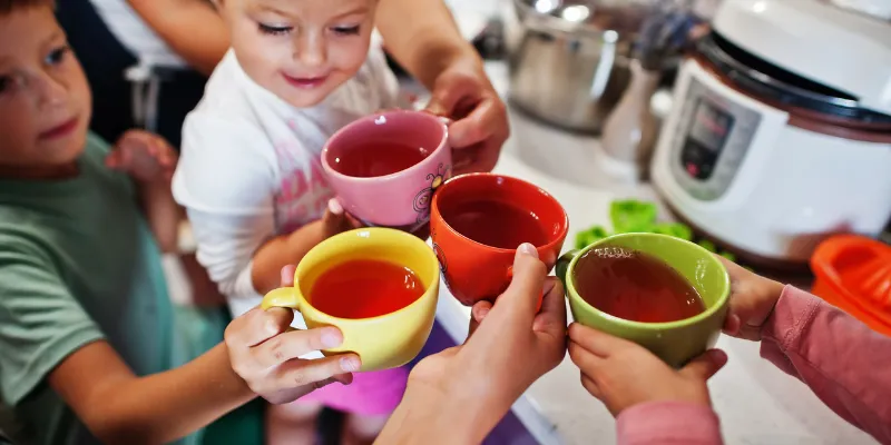 A mother and her young children clinking colorful ceramic mugs filled with a dark fruit drink together in a bright kitchen.