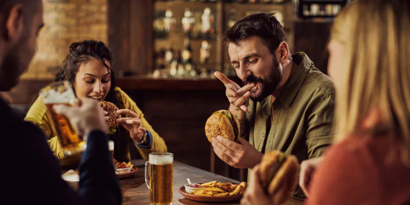 A group of friends laughing and talking while sharing a meal of pizza and burgers with glasses of beer at a busy pub.