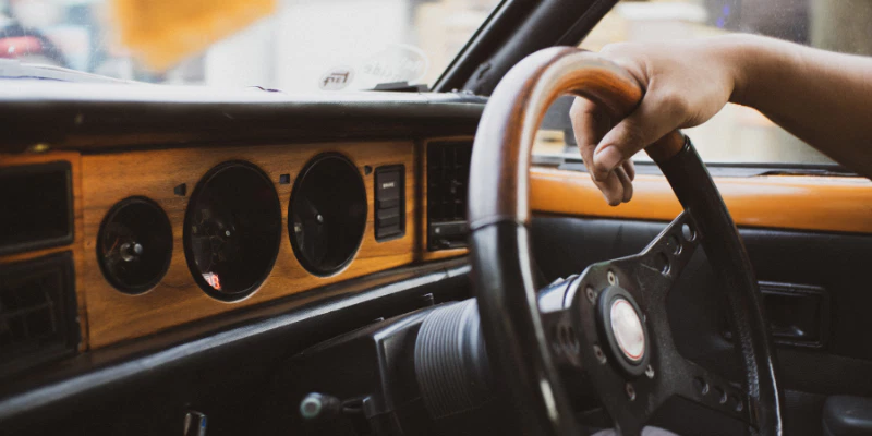 A close-up view of a person's hand resting on the wooden steering wheel of a vintage car with a classic dashboard.