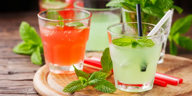 Several vibrant red and green chilled cocktails served in glasses with fresh mint garnishes and straws on a wooden serving board.