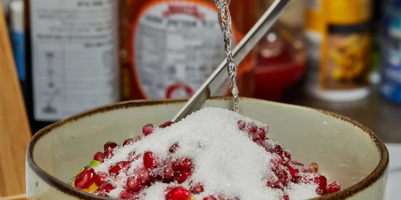 A close-up view of a silver spoon pouring clear liquid simple syrup over fresh pomegranate seeds and sugar in a ceramic bowl.