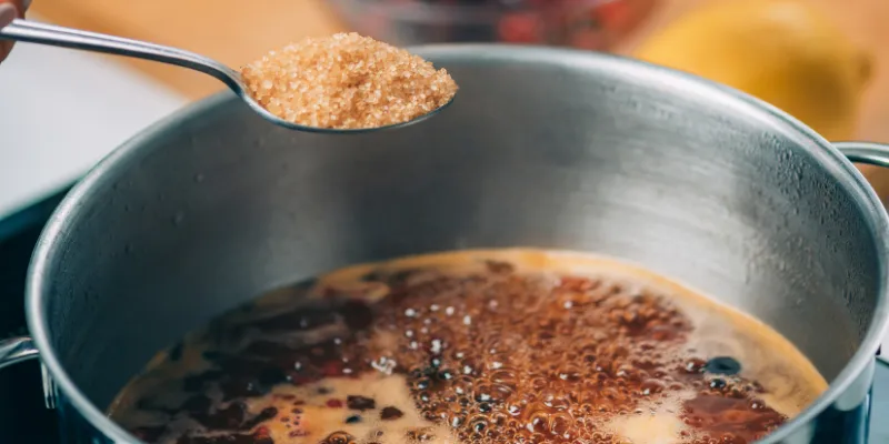 A close-up of a person using a silver spoon to add golden granulated sugar into a stainless steel pot of simmering liquid on a stovetop.