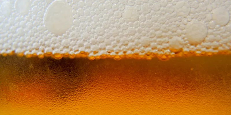 A macro photograph showing the fine, white bubbles of beer foam against a golden, carbonated liquid background.