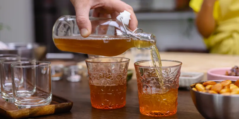 Golden simple syrup being poured from a clear glass swing-top bottle into small decorative glasses on a wooden table.