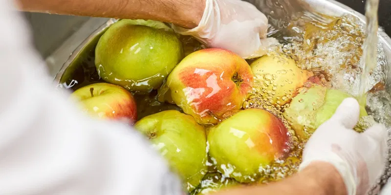 Hands wearing gloves washing a bowl of green and red apples under running water.