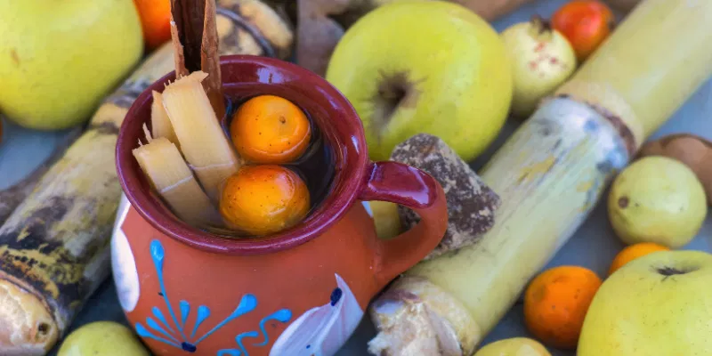 A traditional Mexican clay mug filled with fermented drink, garnished with sugarcane sticks and fruit, surrounded by cinnamon and tejocotes.