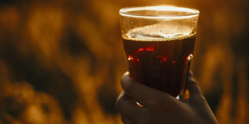 A hand holding a glass of dark, amber-colored sarsaparilla drink against a golden, sunlit background.