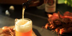 A hand pouring a golden, carbonated beer from a bottle into a glass mug on a dark tabletop.