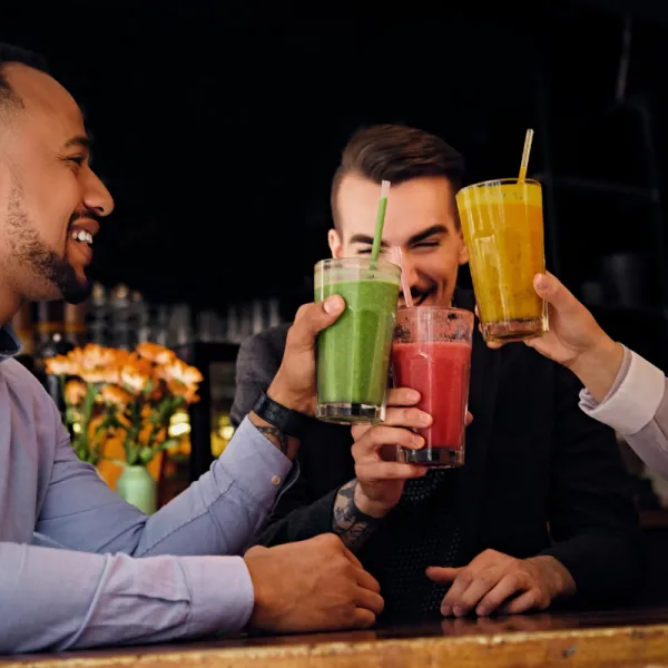 A cheerful group of friends sitting at a bar table, clinking glasses filled with green, red, and yellow non-alcoholic smoothies and juices.