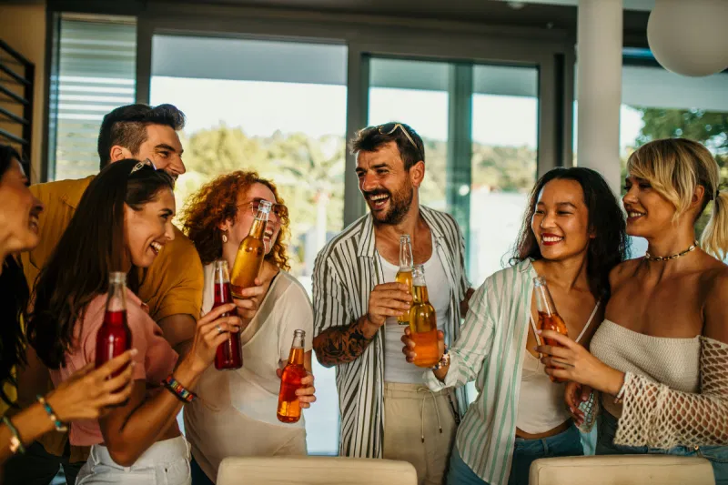 A group of cheerful friends standing indoors at a house party, talking and laughing while holding various bottles of non-alcoholic beverages.
