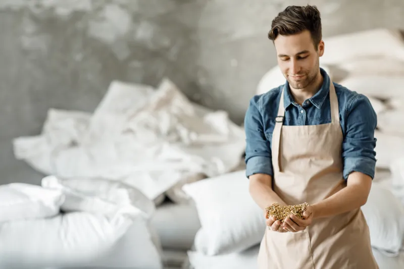 Jonas Leroy, the founder of AlcoFreeBrew, wearing a beige apron and smiling while holding a handful of raw grain malt in a brewery setting.