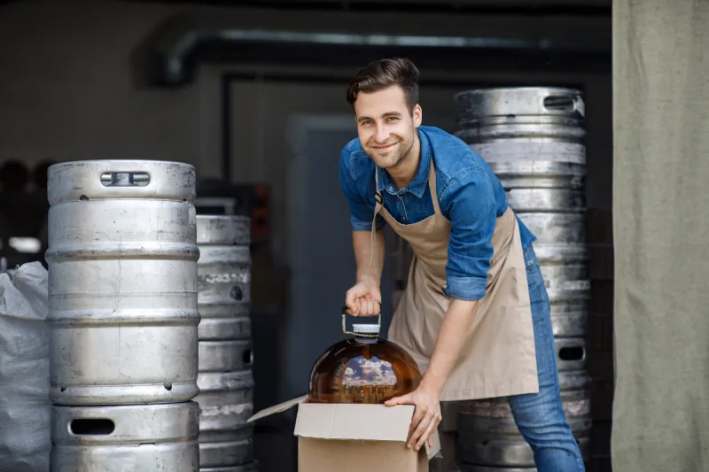 Jonas Leroy crouching down to unpack a large glass carboy (demijohn) fermentation vessel from a cardboard box, looking ready to start a new batch.