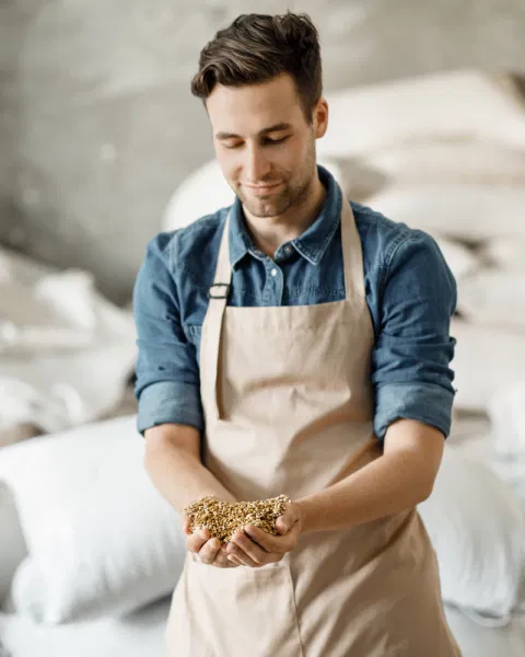 A portrait of Jonas Leroy, the website editor and head brewer, wearing a blue denim shirt and a beige apron, looking down at a handful of malt grains in his hands with a smile.
