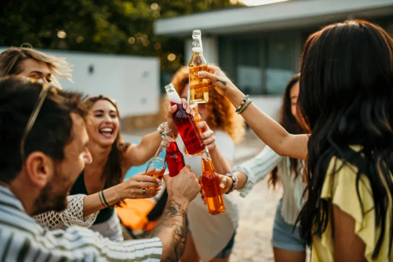 A close-up shot of diverse friends laughing enthusiastically while clinking bottles of red and yellow non-alcoholic drinks together.