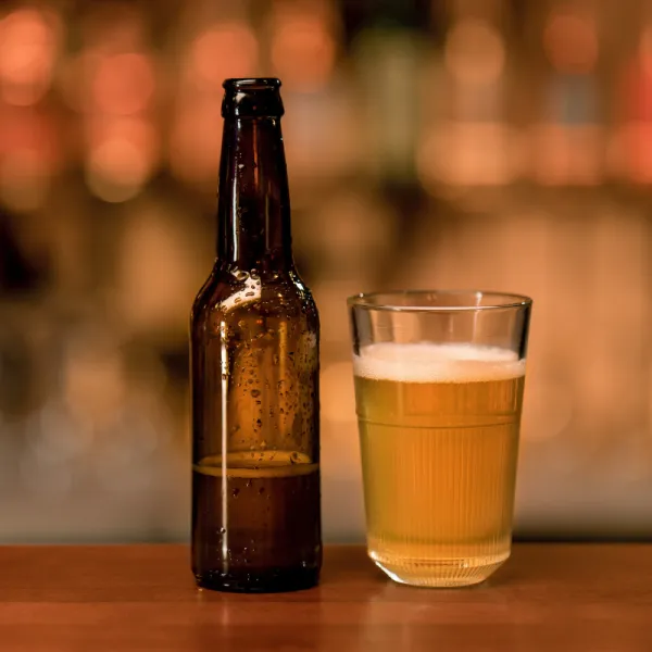 A brown glass beer bottle standing next to a half-pint glass filled with golden non-alcoholic beer on a wooden bar top with a blurred warm background.