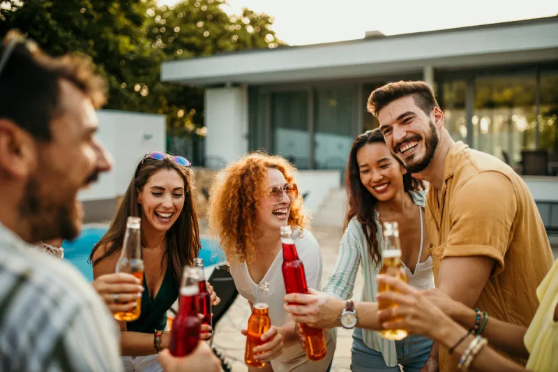 A group of friends raising colorful glass bottles for a toast during a sunny outdoor party, with a swimming pool visible in the background.