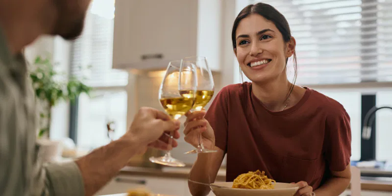 A happy woman in a red shirt toasting with a glass of white zero-proof wine during a pasta lunch at home.