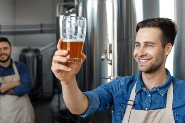A smiling brewer in a beige apron holding up a glass of amber beer to the light to check for clarity, with stainless steel fermentation tanks in the background.